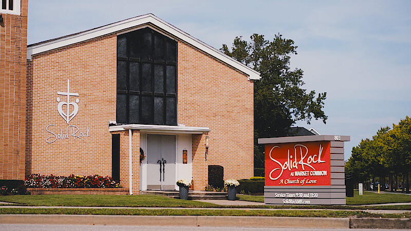 Brick church building with large black window, cross and heart logo, and "Solid Rock" sign out front. Well-maintained lawn, flowers, and trees in a suburban environment under a clear sky.