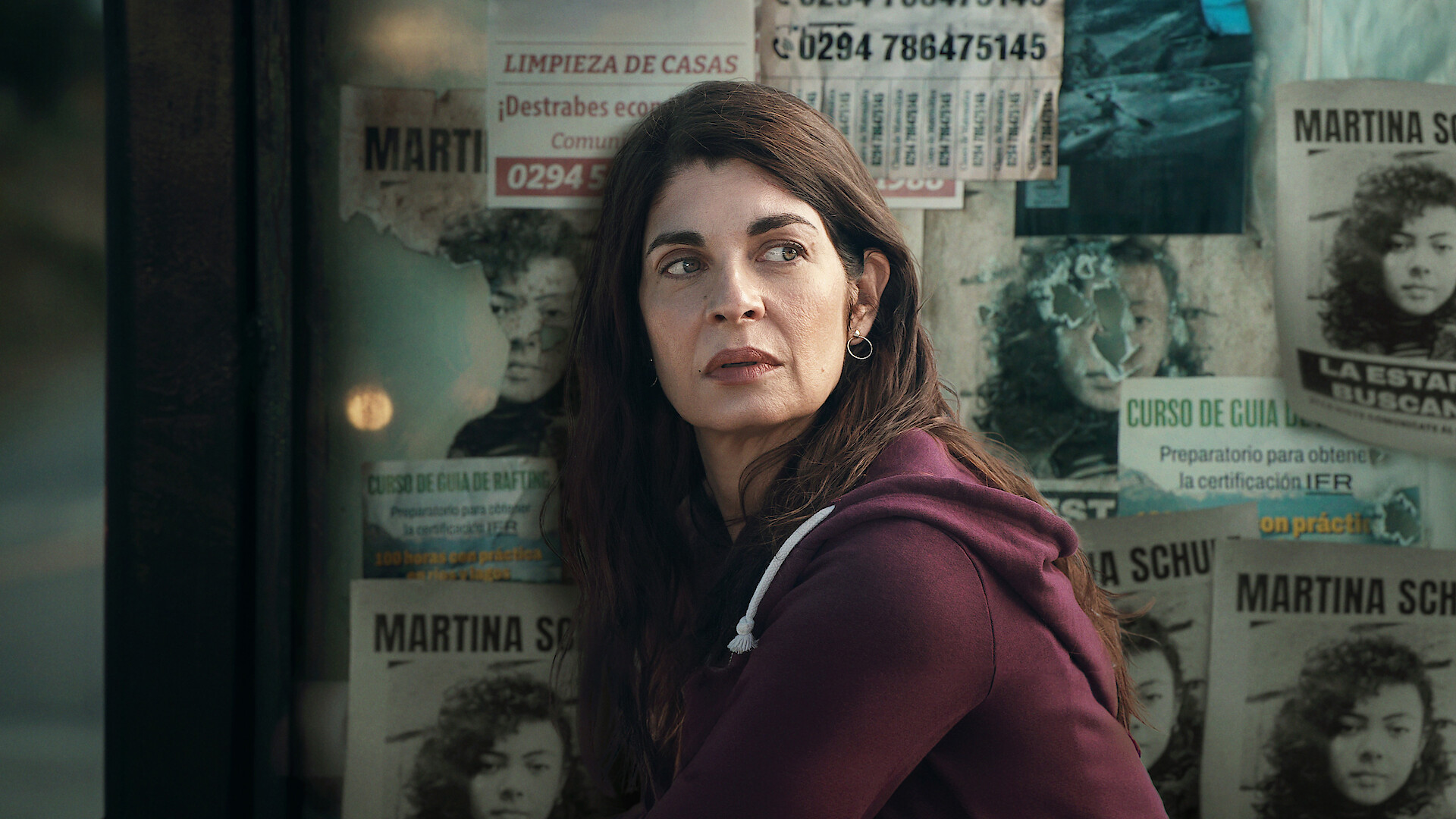 Atrapados key art - A woman looking pensive in front of a wall covered in wanted posters. 