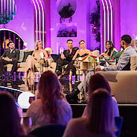 A group of people sit on stage in a brightly lit talk show set with purple lighting, decorative plants, and a live studio audience watching.