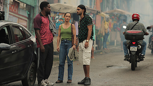 Three people stand talking beside a black car on a busy street with market stalls, colorful buildings, and a person riding a scooter in the background. The scene is urban, vibrant, and lively.