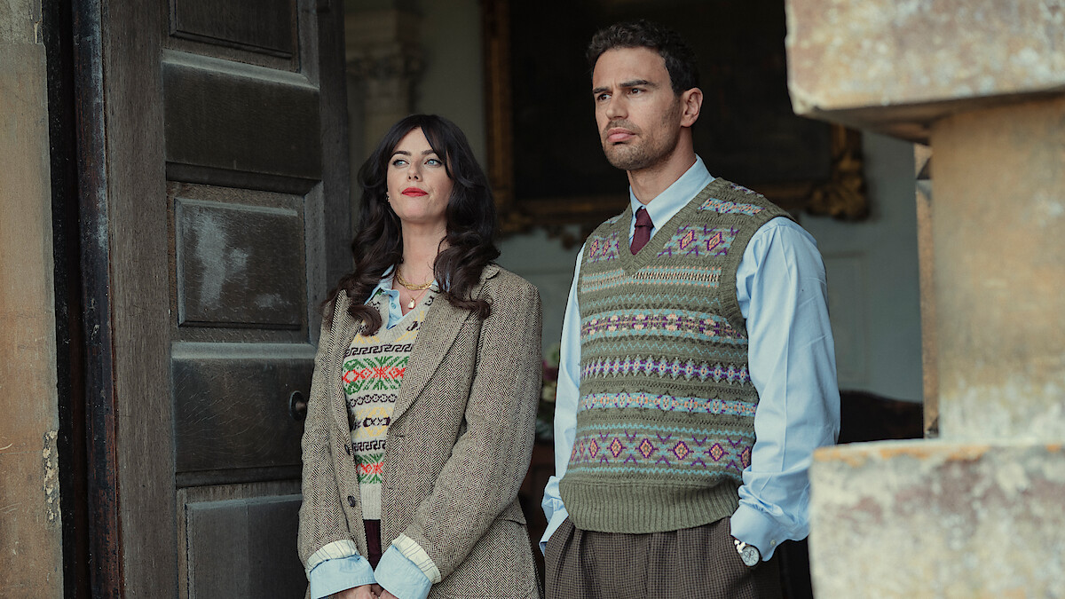 A woman and man stand side by side in vintage-style clothing, both wearing patterned sweater vests and shirts, in a dim, old-fashioned setting with stone and wooden details, evoking a historical or academic atmosphere.