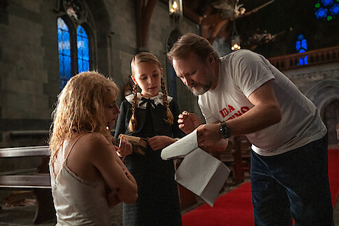 A man gives instructions to two women, one seated and distraught, the other in a school uniform, inside a dimly lit church with stained glass windows, wooden pews, and a serious, contemplative atmosphere.
