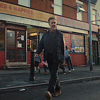 A man in a dark suit walks confidently on a city sidewalk in front of a shop with children playing nearby; the setting features brick buildings and urban street elements in the background.