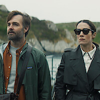 Will Forte as Gilbert Power and Siobhán Cullen as Dove stand together with mountains and water in the background in 'Bodkin.'