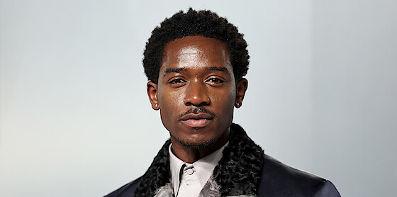 A well-dressed man with short curly hair and earrings standing in front of a plain, light-colored background, photographed in a studio setting with soft lighting.