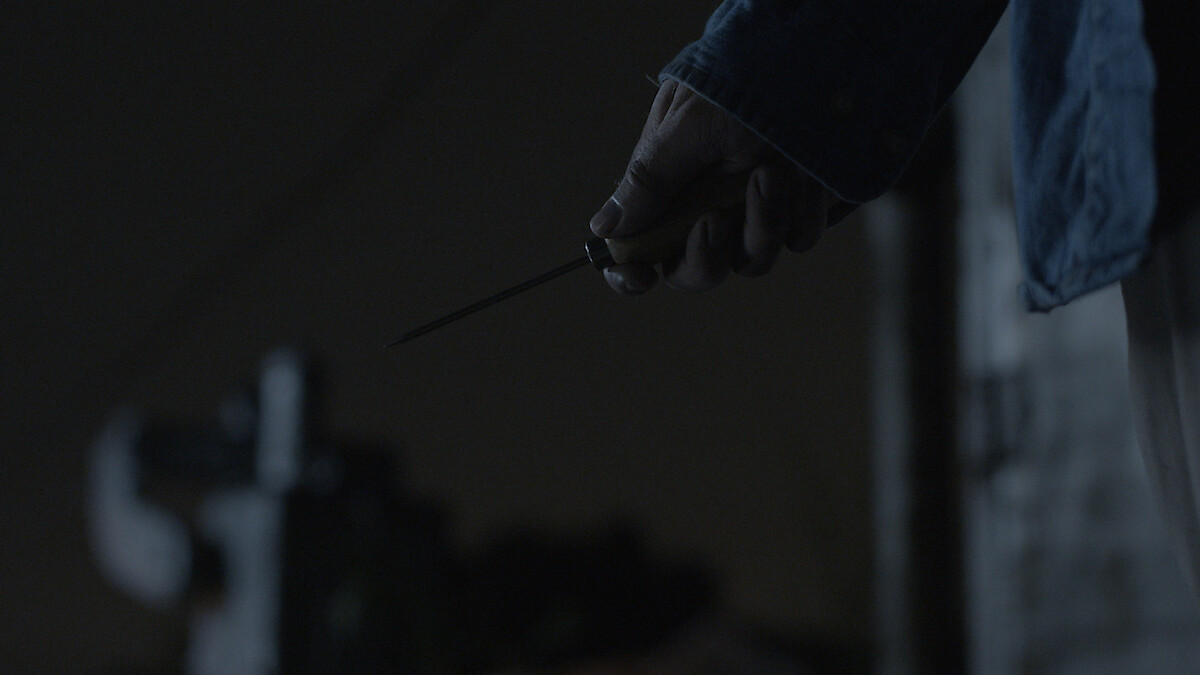 A person holding a knife in their right hand at night, wearing a dark blue jacket. The background is dimly lit, creating a suspenseful or tense atmosphere.
