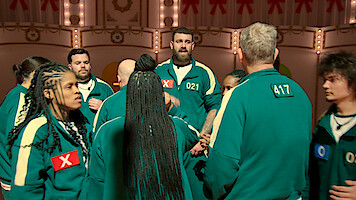 A group of contestants in green tracksuits stand in a lit game arena, some marked with Xs, gathered in a tense huddle, suggesting anticipation and competition in a high-stakes environment.