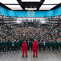 Hundreds of people in green tracksuits stand in rows inside a large, brightly lit room with bunk beds, facing two guards in pink uniforms. 