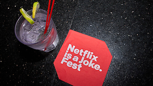 A cocktail with lime slices and two red straws sits on a black granite surface next to a red napkin that reads "Netflix is a Joke Fest" in bold white letters.