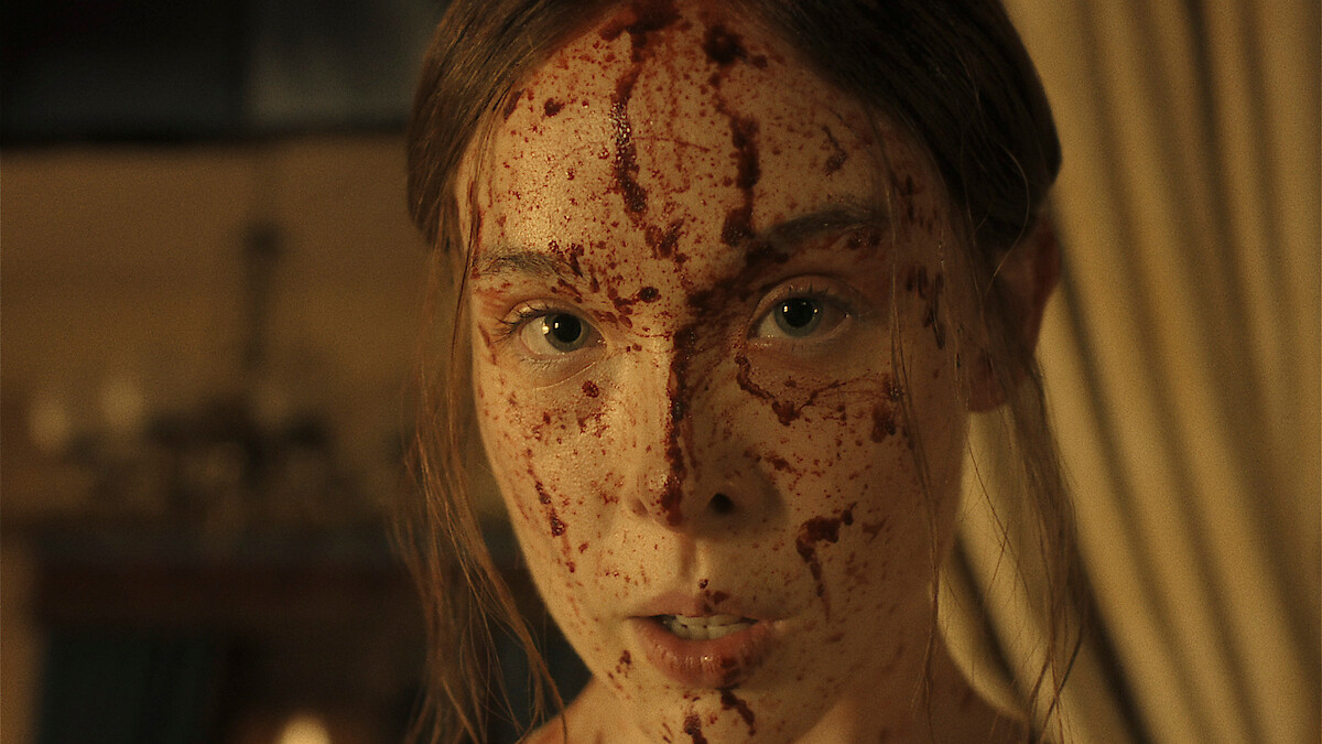 A woman with blood splattered on her face stands indoors in a dimly lit room with curtains and a chandelier visible in the background, looking directly at the camera with an intense expression.