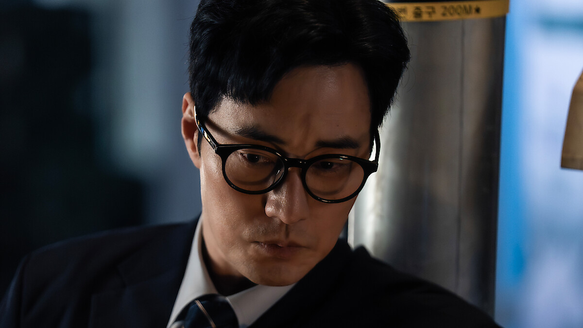 A man in glasses and a suit looks down thoughtfully in a dimly lit indoor setting, near a metal pillar and soft blue background, suggesting a serious or contemplative mood.