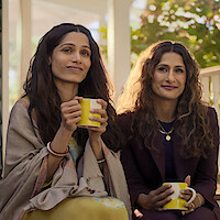 Two women sitting on a porch, holding yellow mugs, dressed in cozy and semi-formal clothes, smiling and enjoying a peaceful outdoor moment with sunlight and greenery in the background.
