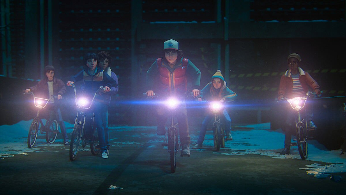 Group of kids riding bikes at night, headlights on, in a dimly lit industrial or urban setting with some patches of snow on the ground, wearing winter clothing and looking determined.