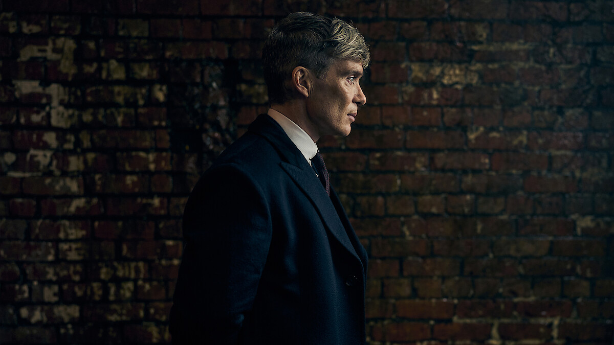 A man in a dark overcoat stands in profile against a worn brick wall in a dimly lit setting, creating a moody and introspective atmosphere.