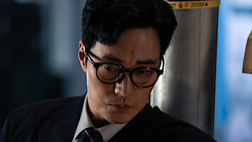 A man in glasses and a suit looks down thoughtfully in a dimly lit indoor setting, near a metal pillar and soft blue background, suggesting a serious or contemplative mood.