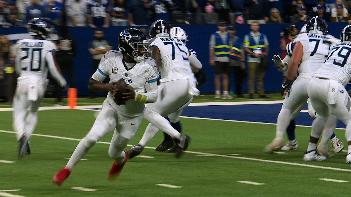 Football quarterback in white uniform running with the ball on a green field during a game, with teammates blocking and stadium spectators in the background.