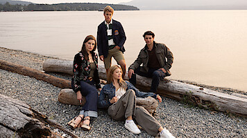 Four young adults posing on driftwood logs at a rocky lakeshore with calm water and a distant treeline in the background, wearing casual clothing and relaxed expressions in an outdoor setting.