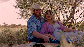 Smiling couple sitting on a blanket outdoors in a grassy field, with trees in the background, wearing casual clothes and cowboy hat, enjoying a relaxed moment in nature during daylight.