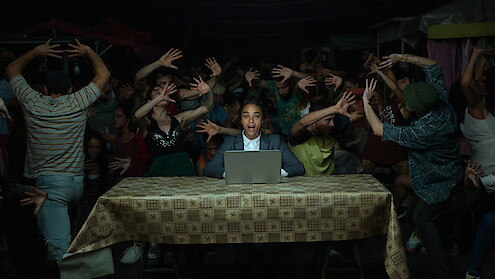 Rita (Zoe Saldaña) sits at a table behind a laptop, as people behind her dance wildly.