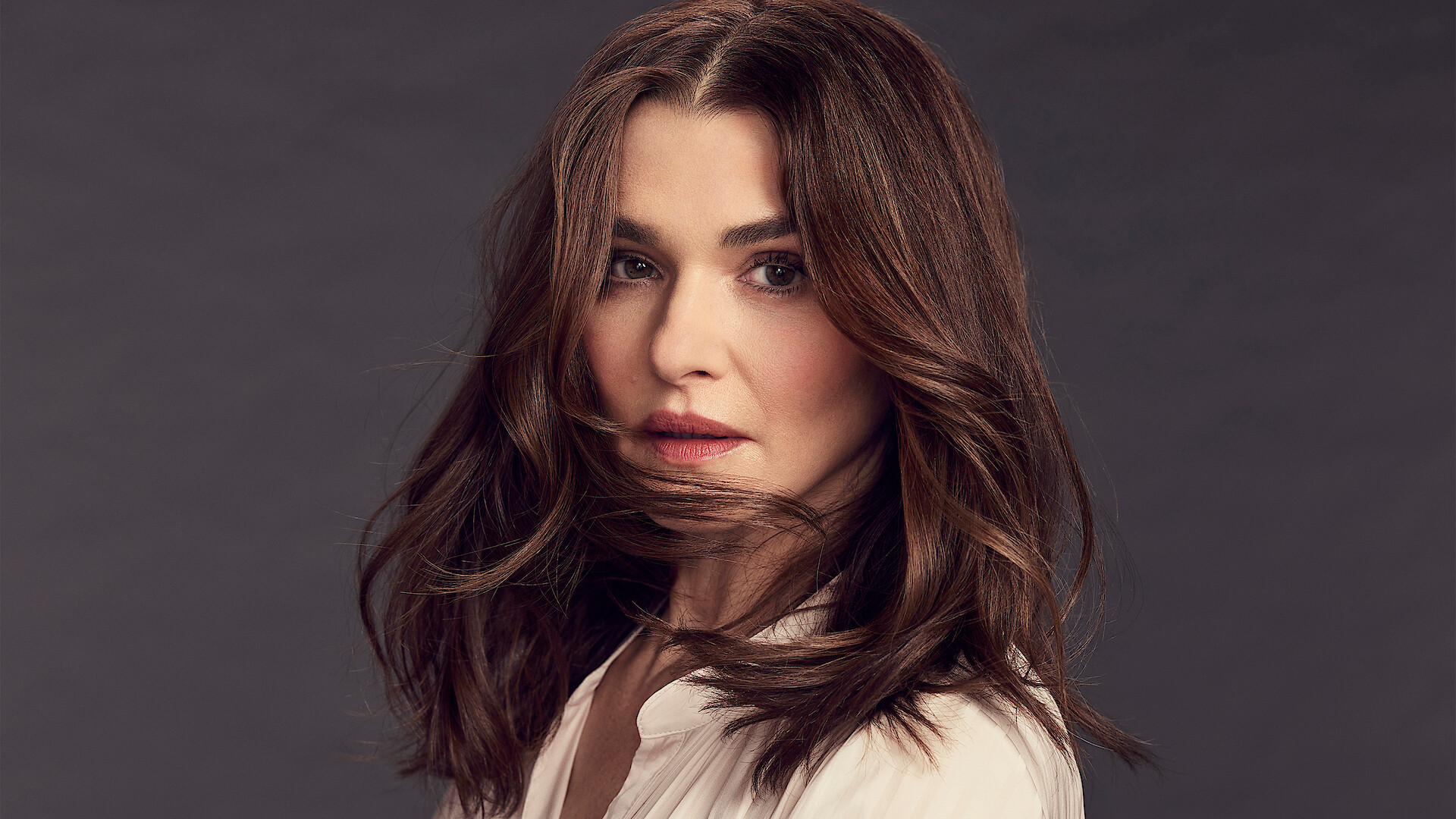 Rachel Weisz stands in front of a grey background wearing a white blouse, caught mid head turn with a clump of hair wrapping around her chin while looking to camera.