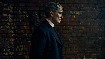 A man in a dark overcoat stands in profile against a worn brick wall in a dimly lit setting, creating a moody and introspective atmosphere.