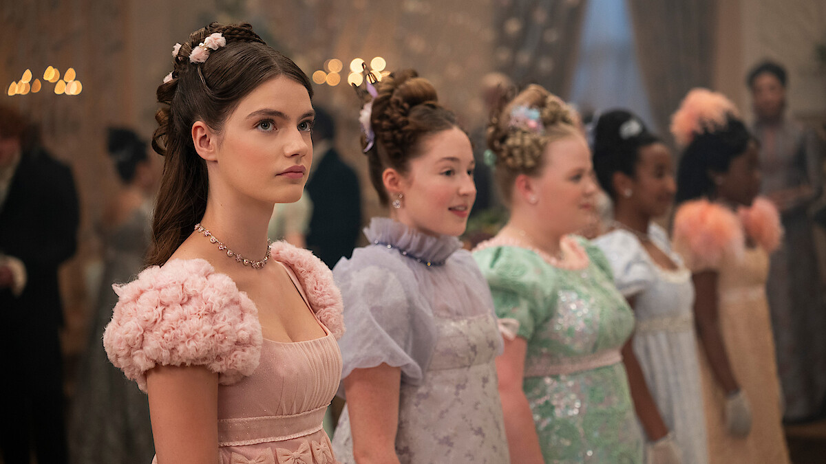 Young women dressed in elegant, pastel-colored Regency-era gowns stand in a row at a formal ball, adorned with floral hair accessories, with candlelight and guests blurred in the ornate, softly lit background.