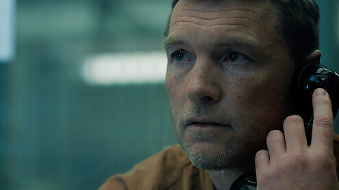 Man with short hair and stubble sits indoors, holding a black phone receiver to his ear, looking serious. The background is blurred, suggesting he is in a visitation booth or interrogation room, likely in a prison or secure facility.