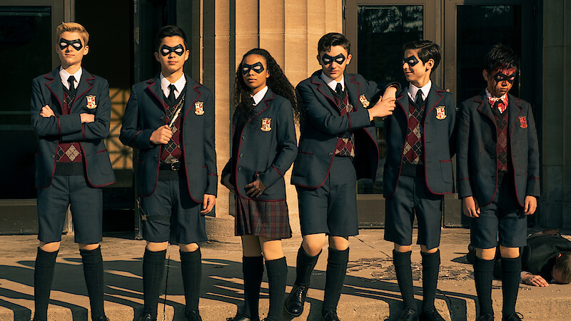 Six children in matching school uniforms and black eye masks stand in a row outside a building with large columns, in sunlight, appearing serious or ready for action. Another person is lying on the ground in the background.