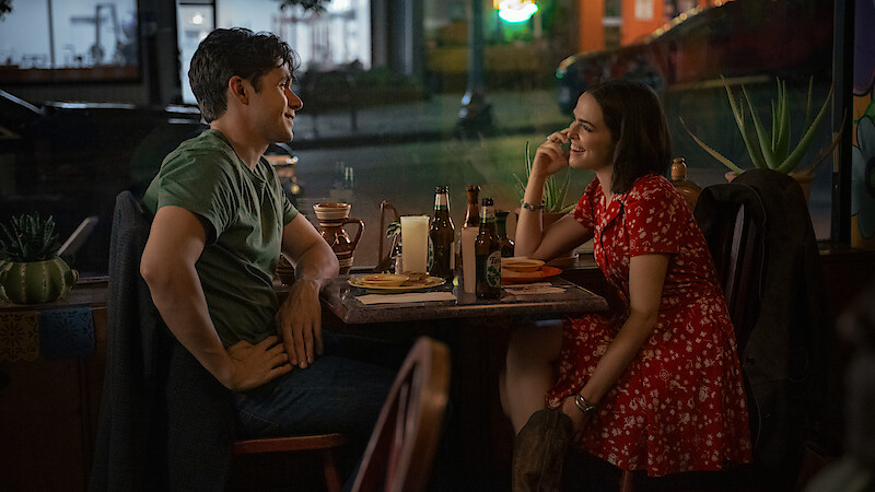 A man and woman sit at a restaurant table at night, smiling and talking over drinks and food, creating a warm, intimate, and relaxed atmosphere with streetlights visible outside the window.