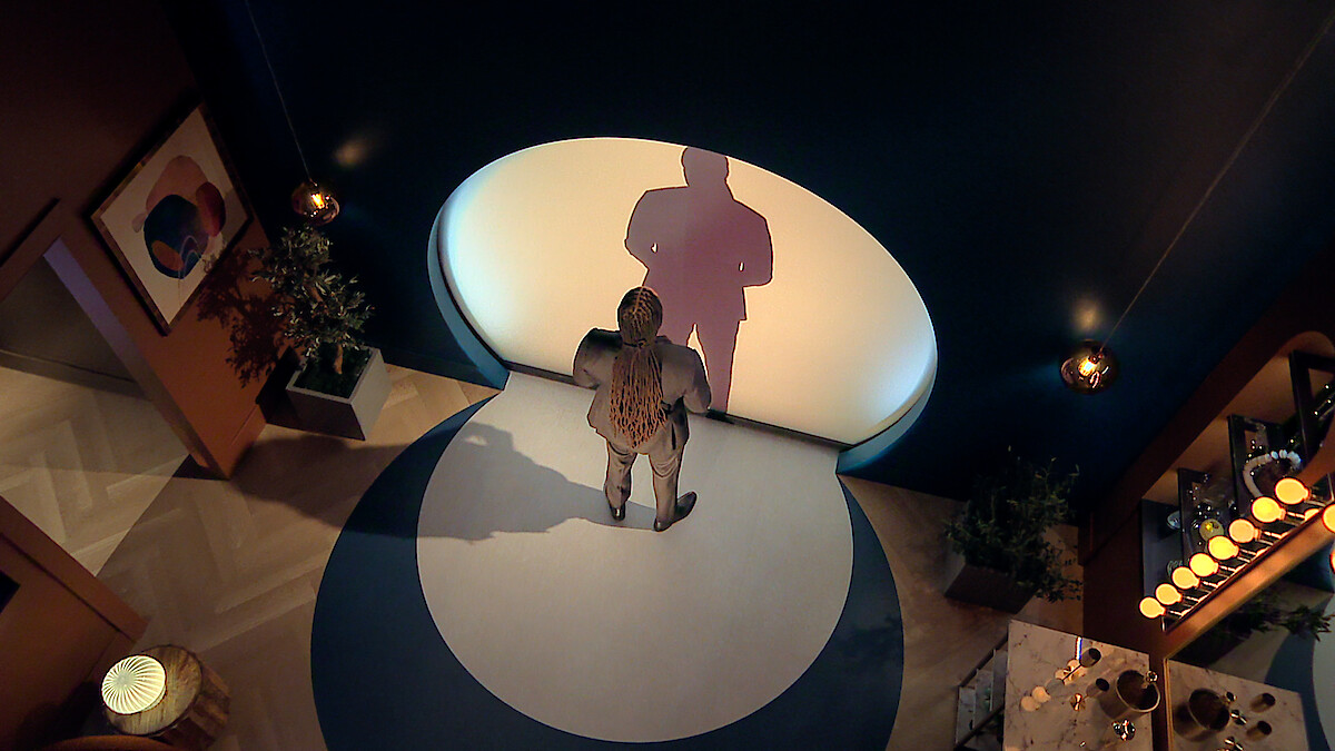 Dr Vic St John standing alone in a modern, dimly lit room with blue walls, casting a large shadow onto a circular white light on the wall. Decorative plants, artwork, and a set dining table create a stylish, intimate environment.