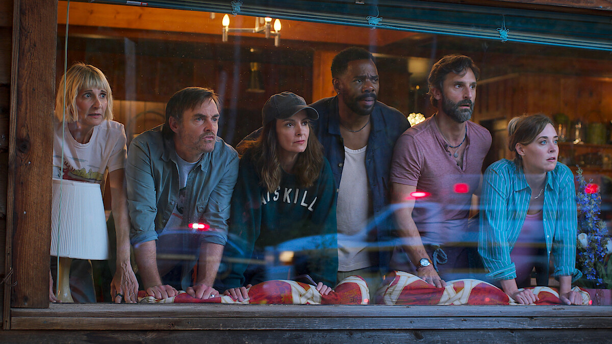 Six people lean on a windowsill inside a rustic wooden building, looking outside with serious, focused expressions. The setting has warm lighting and cozy decor, with reflections of lights and cars visible on the glass.