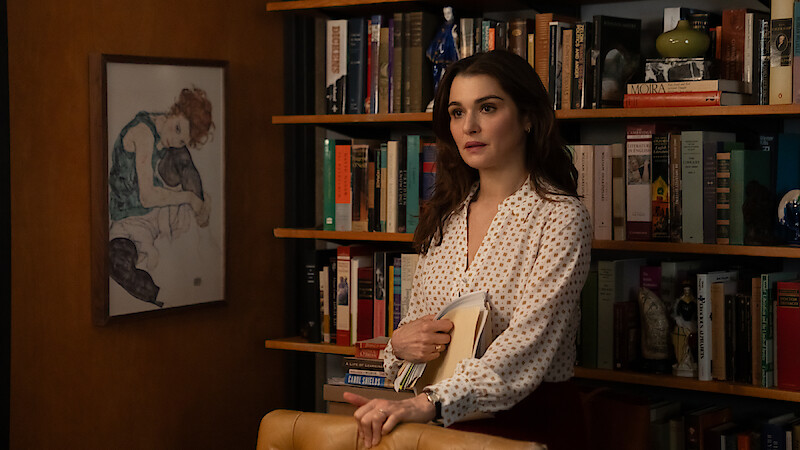 Rachel Weisz as The Protagonist  in a white blouse stands in front of a bookshelf filled with books and decor, holding folders. A framed artwork hangs on the wall, creating a cozy, intellectual office or library setting.