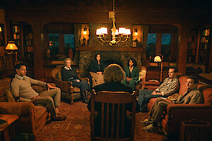 Seven people sit in a cozy, warmly lit cabin living room arranged in a circle, suggesting a group discussion or therapy session, with a rustic fireplace, wooden beams, and a calm, contemplative atmosphere.