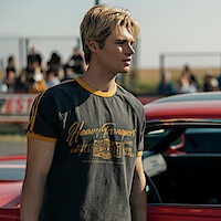 Young man with blond hair wearing a "Heavy Transport" t-shirt stands next to a red car at an outdoor event with spectators and a metal fence visible in the background.