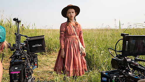 Young girl in a vintage dress and hat stands in a grassy field holding a book, surrounded by film cameras, during an outdoor movie or TV production.