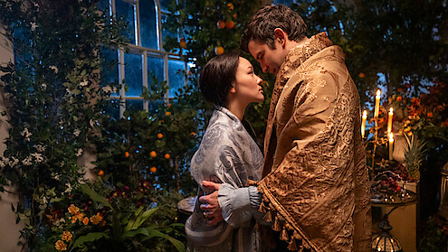 A man and woman in ornate robes share an intimate moment in a lush, candlelit indoor garden, surrounded by plants, flowers, and fruits near a large window at night.