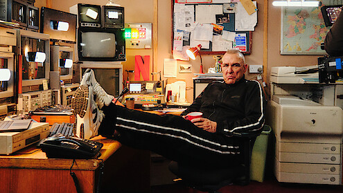 Vinnie Jones in a tracksuit relaxes with feet on desk in a cluttered control room, surrounded by monitors, electronics, bulletin board, and office equipment, holding a red cup.