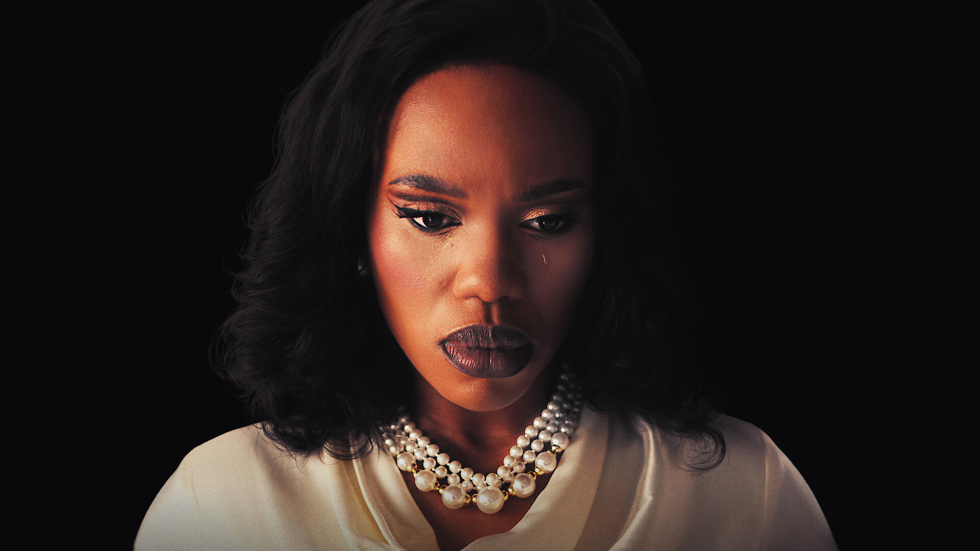Woman with dark hair and makeup wearing a white blouse and layered pearl necklaces, looking down with a serious expression against a dark background.