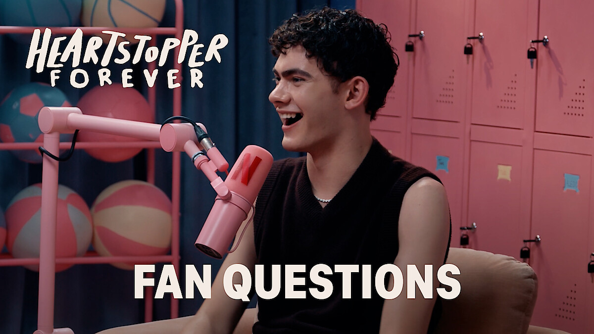 Joe Locke smiles and talks into a pink microphone in a colorful room with lockers and pastel basketballs, with "Heartstopper Forever" and "Fan Questions" text on screen, suggesting a fan Q&A interview setting.