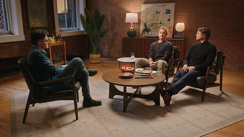 Three people have a conversation seated in a cozy, warmly lit living room with modern decor, including a coffee table, books, fruit bowl, plants, and large abstract art on the brick wall.