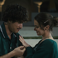 A young man and woman share an intimate moment seated together at night; the man gently touches the woman's hand as she smiles and admires a ring, suggesting a romantic or engagement setting.