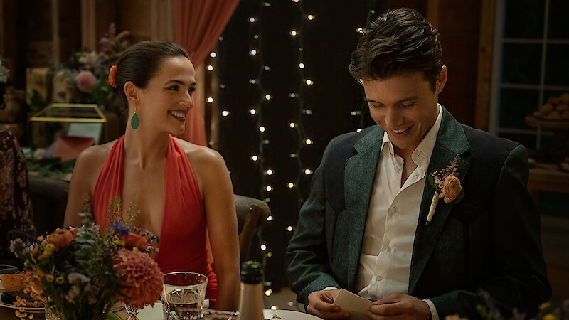 A woman in a red dress and a man in a suit sit at a decorated dinner table, smiling and reading a note, with warm string lights and floral arrangements in a cozy, intimate indoor setting.
