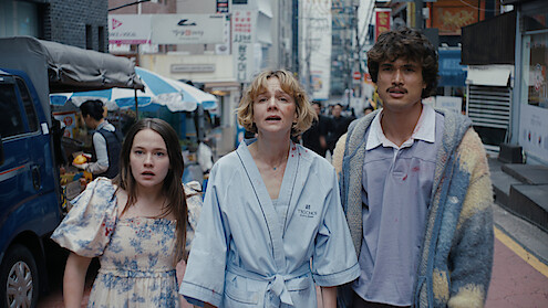 Cailee Spaeny as Ashley Miller, Carey Mulligan as Lindsay Crane-Martin, and Charles Melton as Austin Davis in casual clothing stand on a busy city street, looking upward with surprised expressions, surrounded by vendors, shops, and tall buildings.