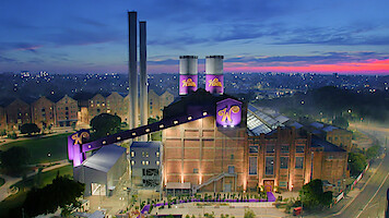 Large whimsical chocolate factory with purple “W” logos, brick exterior, and tall chimneys, set in a city at dusk. Brightly lit, surrounded by greenery and roads, with apartments in the background under a colorful sky.