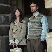 A woman and man stand in a doorway wearing vintage-style clothing with patterned sweaters; the setting appears formal and indoors, with a sophisticated, historic atmosphere.