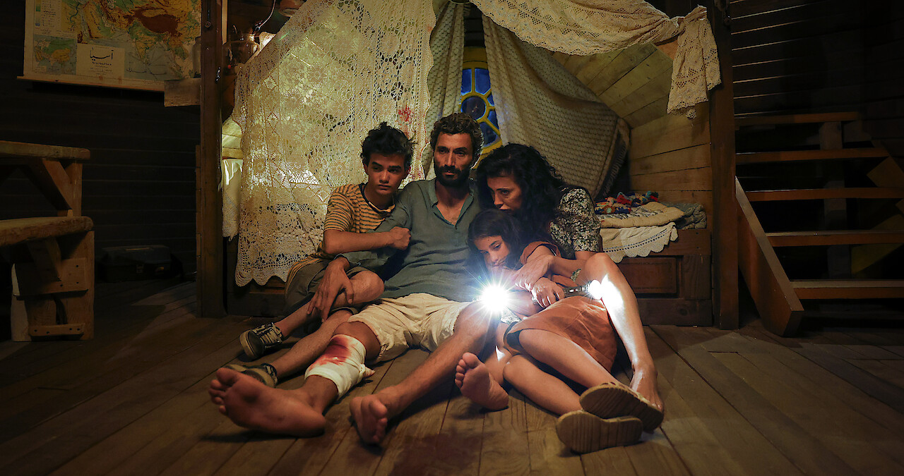 Riman Al Rafeea, Ziad Bakri, Zain Al Rafeea, and Nadine Labaki huddle together in a small room on a wood floor in the dark in an image from the 2025 film 'The Sand Castle.'