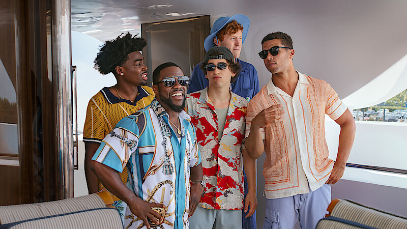 Five men in colorful summer shirts are standing and talking on a luxury boat deck, smiling and laughing together in a relaxed, sunny outdoor setting.