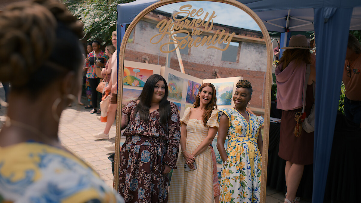 Three women smiling and posing together in front of a decorative mirror at an outdoor event, with other people and a tent in the background. The mirror has the words "See Yourself in Serenity" written on it.