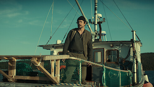 A man wearing a beanie stands on the deck of a fishing boat surrounded by nets and wooden railings, with the sea and distant land visible under a clear sky.