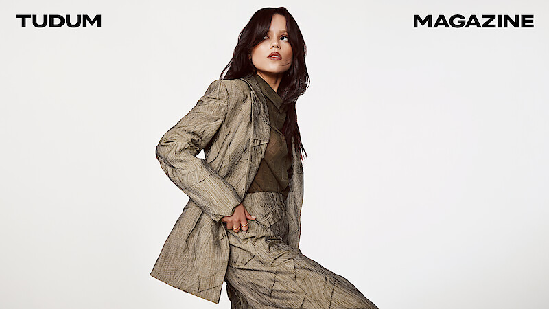 Jenna Ortega in a taupe textured suit with sheer blouse looks to the side, gaze locked on something in the distance with her hair down.
